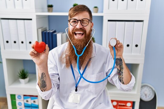 Redhead man with long beard wearing doctor uniform holding heart and stethoscope winking looking at the camera with sexy expression, cheerful and happy face.