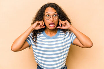 Young hispanic woman isolated on beige background covering ears with fingers, stressed and desperate by a loudly ambient.