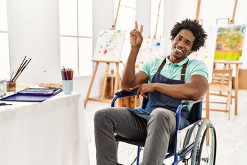Young african american artist man sitting on wheelchair at art studio smiling with happy face winking at the camera doing victory sign. number two.