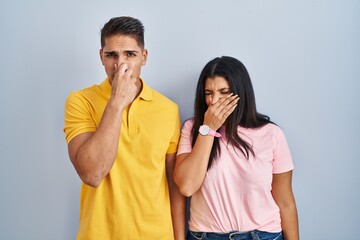 Young couple standing over isolated background smelling something stinky and disgusting,...