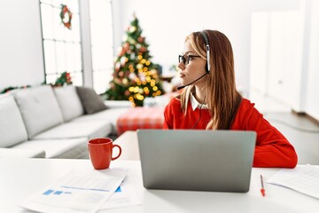 Young caucasian girl sitting on the table working using laptop by christmas tree looking to side, relax profile pose with natural face with confident smile.