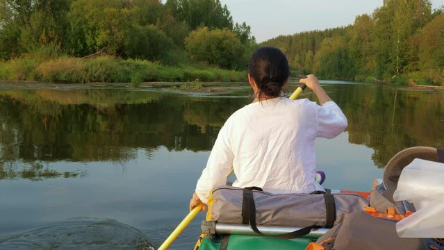 Girl is sailing on boat on river surrounded by forest in evening. Asian woman rowing paddle on catamaran. Female boater, tourist. Sunset, summer. Sportive lifestyle. Back view. Active vacation