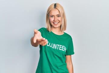 Beautiful blonde woman wearing volunteer t shirt smiling cheerful offering palm hand giving assistance and acceptance.