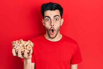 Young hispanic man holding peanuts scared and amazed with open mouth for surprise, disbelief face