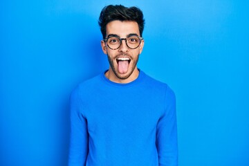 Young hispanic man wearing casual clothes and glasses sticking tongue out happy with funny expression. emotion concept.
