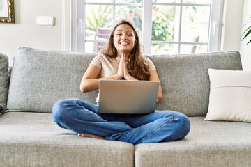 Beautiful young brunette woman sitting on the sofa using computer laptop at home praying with hands together asking for forgiveness smiling confident.