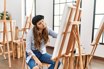 Young hispanic artist woman painting on canvas at art studio looking sleepy and tired, exhausted...