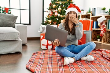 Young latin woman using laptop sitting by christmas tree shocked covering mouth with hands for mistake. secret concept.