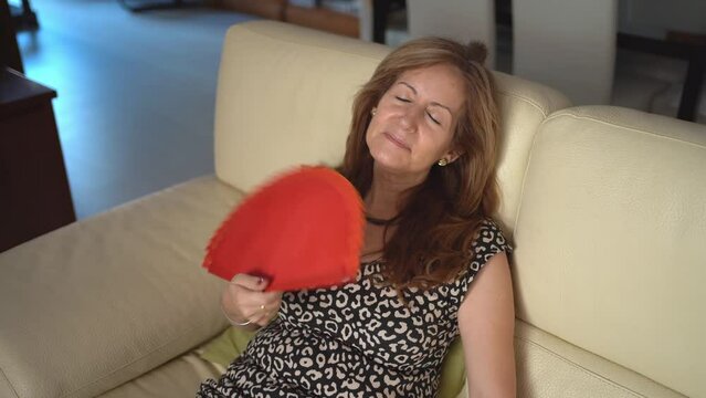 Mature woman sitting on a sofa and giving herself fresh air with a fan through the high heat of summer.