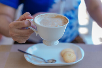 Close up of lady's hands holding white coffee cup with blur cookies at cafe table during coffee break or breakfast in vacations. Traveler woman holding white mug of hot coffee for relaxing on holidays
