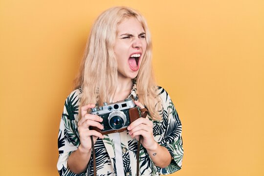 Beautiful caucasian woman with blond hair holding vintage camera angry and mad screaming frustrated and furious, shouting with anger. rage and aggressive concept.
