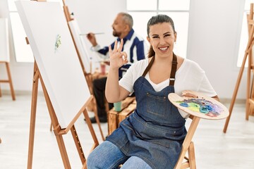 Young artist woman at art studio doing ok sign with fingers, smiling friendly gesturing excellent symbol