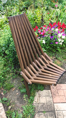 A dark brown wooden folding chair stands on the ground, surrounded by green bushes, flowers and a garden path made of tiles