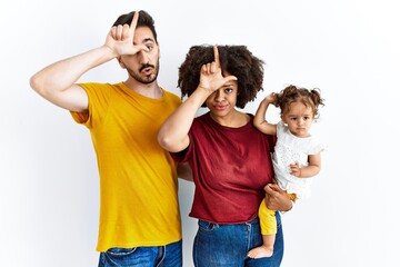 Interracial young family of black mother and hispanic father with daughter making fun of people with fingers on forehead doing loser gesture mocking and insulting.