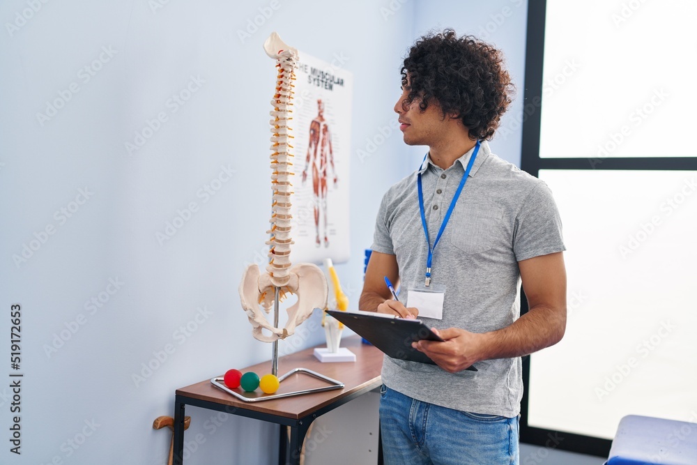 Sticker young hispanic man physiotherapist writing on clipboard at rehab clinic