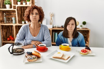Family of mother and down syndrome daughter sitting at home eating breakfast puffing cheeks with funny face. mouth inflated with air, crazy expression.