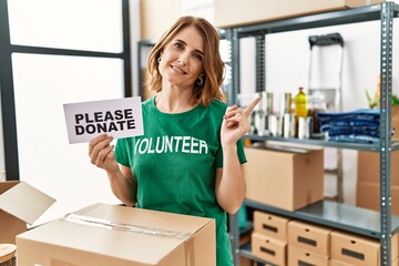 Middle age woman wearing volunteer t shirt holding please donate banner smiling happy pointing with hand and finger to the side