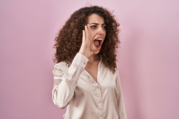 Hispanic woman with curly hair standing over pink background shouting and screaming loud to side with hand on mouth. communication concept.