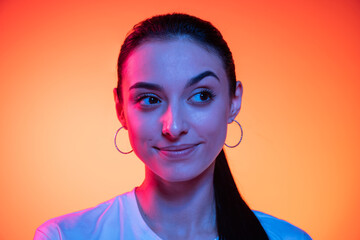 Closeup face of young attractive girl looking at camera isolated on orange color background in neon light. Concept of emotions, facial expressions