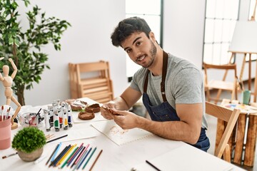 Young hispanic artist man smiling happy modeling pottery at art studio.