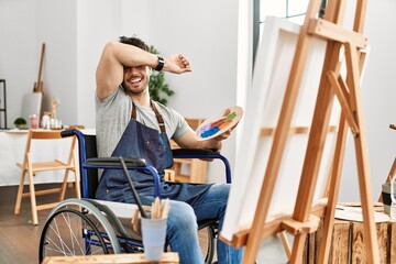 Young hispanic man sitting on wheelchair painting at art studio smiling cheerful playing peek a boo with hands showing face. surprised and exited