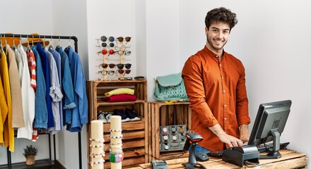 Young hispanic shopkeeper man smiling happy working at clothing store. - obrazy, fototapety, plakaty