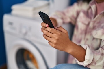 Young african american woman using smartphone waiting for washing machine at laundry