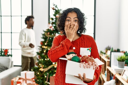 Young hispanic woman standing by christmas tree with decoration laughing and embarrassed giggle covering mouth with hands, gossip and scandal concept