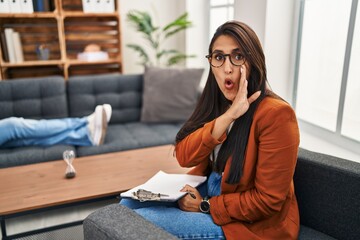 Young hispanic woman working as psychology counselor hand on mouth telling secret rumor, whispering malicious talk conversation