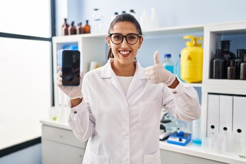 Young hispanic woman working at scientist laboratory with smartphone smiling happy and positive, thumb up doing excellent and approval sign