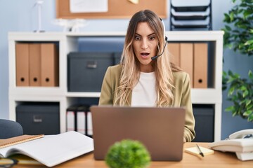 Young hispanic woman wearing call center agent headset working at the office scared and amazed with...