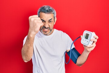 Handsome middle age man with grey hair using blood pressure monitor annoyed and frustrated shouting...