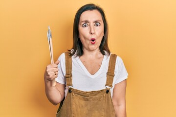 Middle age hispanic woman holding paintbrushes scared and amazed with open mouth for surprise,...