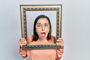 Middle age hispanic woman holding empty frame afraid and shocked with surprise and amazed expression, fear and excited face.