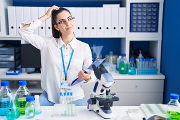 Young brunette woman working at scientist laboratory confuse and wondering about question. uncertain with doubt, thinking with hand on head. pensive concept.