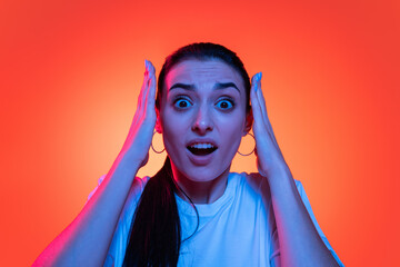 Young emotional stressed girl, student in white t-shirt looking at camera wide open eyes isolated on orange color background in neon light.
