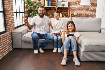 Family of three playing video game sitting on the sofa smiling happy pointing with hand and finger to the side