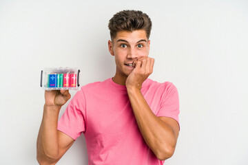 Young caucasian man holding a batteries box isolated on white background biting fingernails,...
