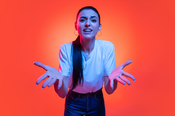 Young emotional girl, student in white t-shirt looking at camera wide open eyes isolated on orange color background in neon light.