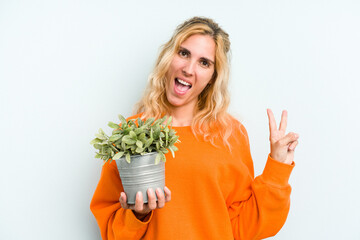 Young caucasian woman holding a plant isolated on blue background joyful and carefree showing a peace symbol with fingers.