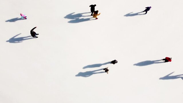 Many people are skating on a white outdoor ice rink in the city on a sunny winter day. Shadows of people skating on the surface of a white ice rink. Aerial drone view. Top view. Lifestyle, sport, rest