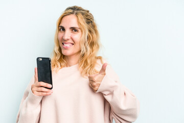 Fototapeta premium Young caucasian woman holding mobile phone isolated on blue background smiling and raising thumb up