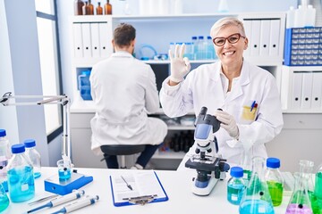Middle age woman working at scientist laboratory doing ok sign with fingers, smiling friendly gesturing excellent symbol