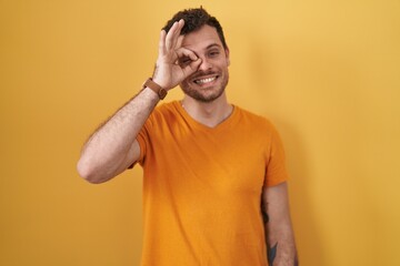 Fototapeta na wymiar Young hispanic man standing over yellow background doing ok gesture with hand smiling, eye looking through fingers with happy face.