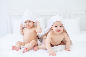 two twin babies in a white towel after bathing in a bathtub on a bright bed at home smiling, the...