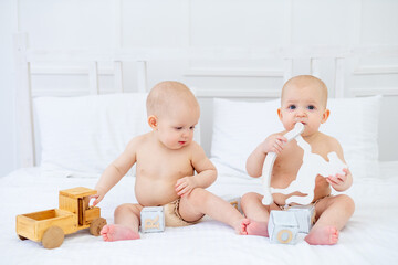 two twin babies girls of six months in diapers on a white cotton bed in a bodysuit on a bed at home playing with wooden toys and smiling