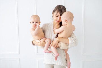 a young mother holds two twin babies in diapers in a bright room at home and smiles, happy...