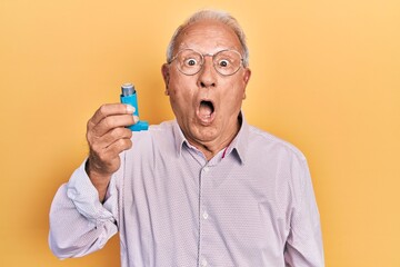 Senior man with grey hair holding medical asthma inhaler scared and amazed with open mouth for...