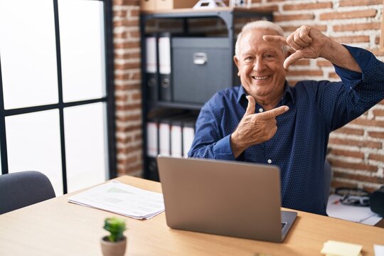 Senior man with grey hair working using computer laptop at the office smiling making frame with hands and fingers with happy face. creativity and photography concept.