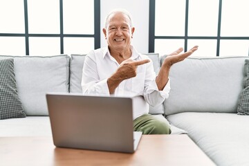 Senior man using laptop at home sitting on the sofa amazed and smiling to the camera while presenting with hand and pointing with finger.
